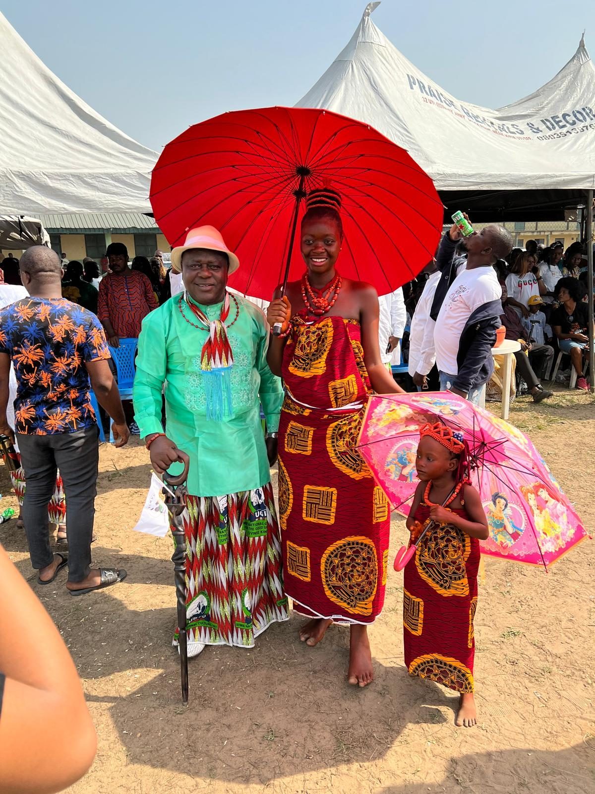 A young woman in a red george wrapper and coral beads under a red ceremonial umbrella, a child beside her dressed the same.